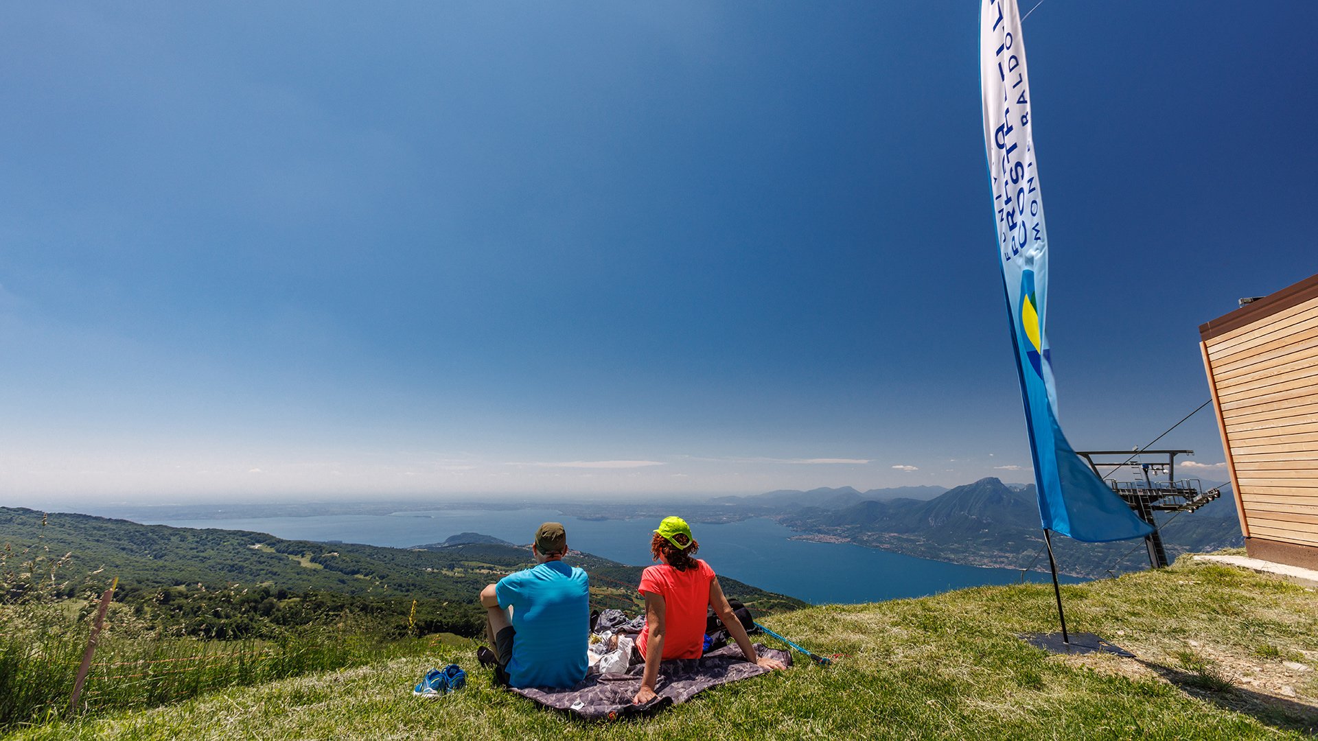 Paar sitzt auf dem Gras des Monte Baldo, mit atemberaubender Aussicht auf den Gardasee und die umliegenden Berge